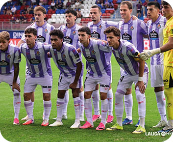 Un grupo de personas vestidas con uniformes deportivos morados y blancos posan juntas en un campo de f tbol antes de un partido. Descripci n generada con IA