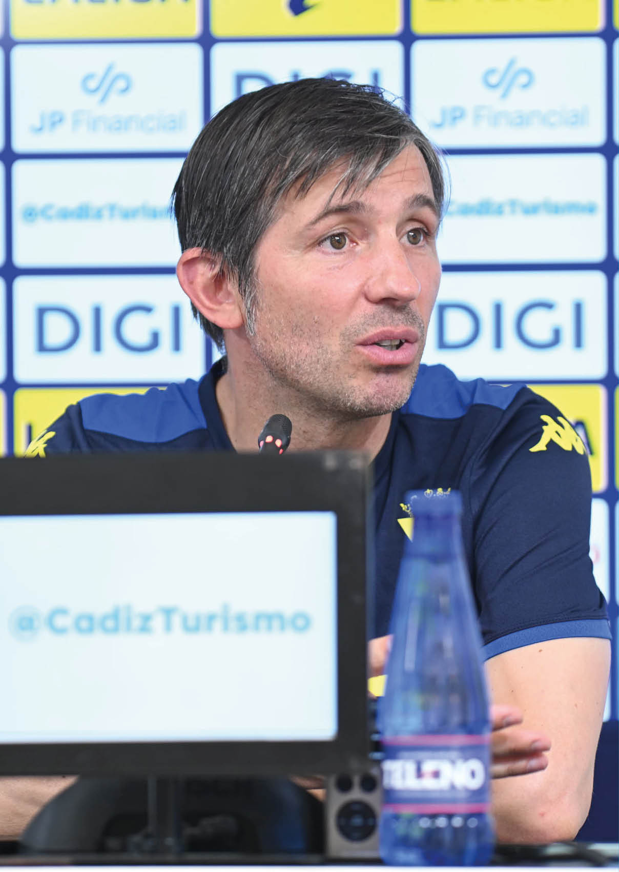 Una persona con camiseta deportiva azul habla frente a un micr fono en una conferencia de prensa con un fondo lleno de logotipos y una botella de agua en primer plano. Descripci n generada con IA