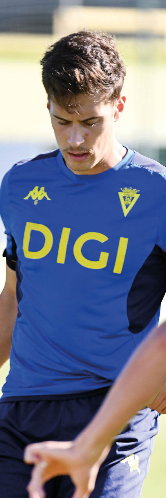 Una persona con camiseta azul y pantalones cortos oscuros est controlando un bal n de f tbol blanco en un campo de c sped.  Descripci n generada con IA