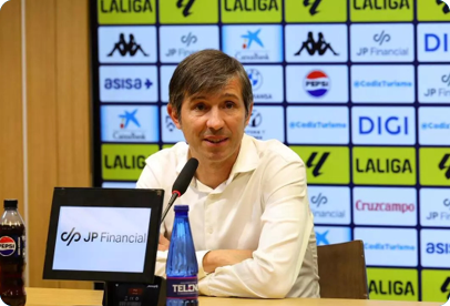 A person in a white shirt is sitting at a table with a microphone, a water bottle, and a screen displaying “JP Financial" in front of a backdrop featuring various logos including LaLiga and Pepsi. Descripci n generada con IA