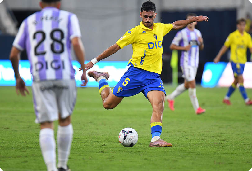 A person in a yellow and blue soccer uniform is kicking a soccer ball on a grass field while other players in white and purple uniforms watch. Descripci n generada con IA