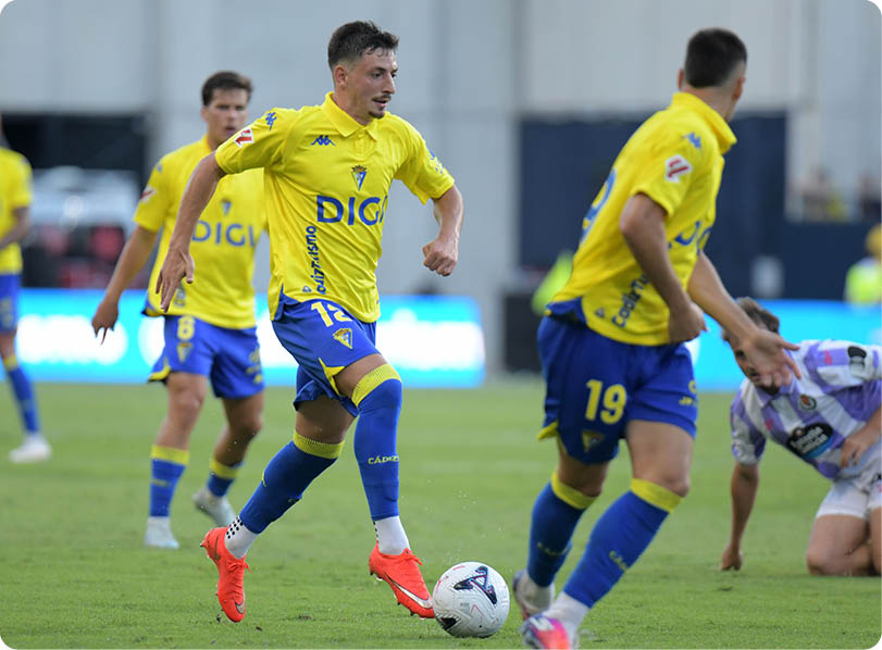 People wearing blue and yellow sports uniforms are playing soccer on a grass field with one person about to kick the ball. Descripci n generada con IA