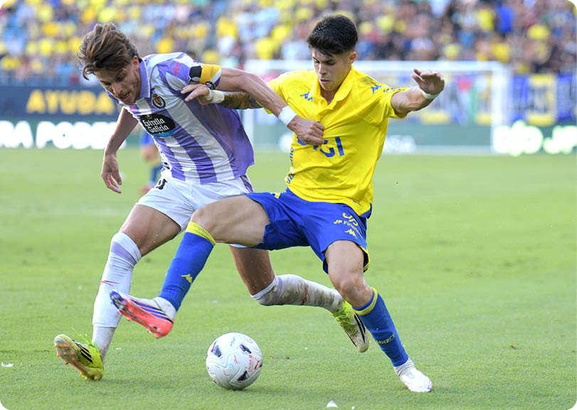Two people in sports uniforms are competing for a soccer ball on a grassy field during a game. Descripci n generada con IA