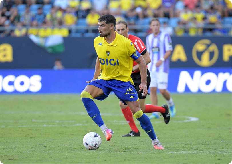 A person wearing a yellow and blue soccer uniform is controlling a soccer ball on a grassy field with other players and a referee in the background. Descripci n generada con IA