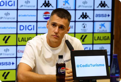 A person with short hair wearing a white t-shirt is sitting at a table with a microphone and a bottle of Pepsi in front of a backdrop covered with various sponsor logos. Descripci n generada con IA