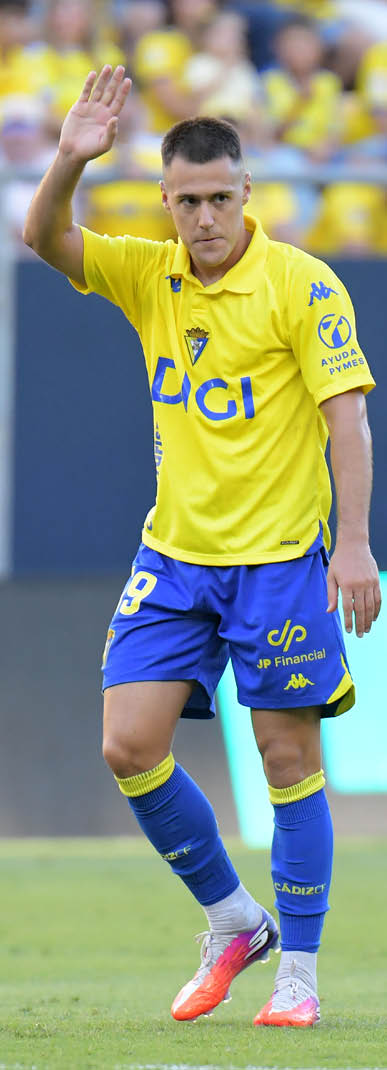 A person wearing a yellow and blue soccer uniform is raising one hand while standing on a soccer field. Descripci n generada con IA
