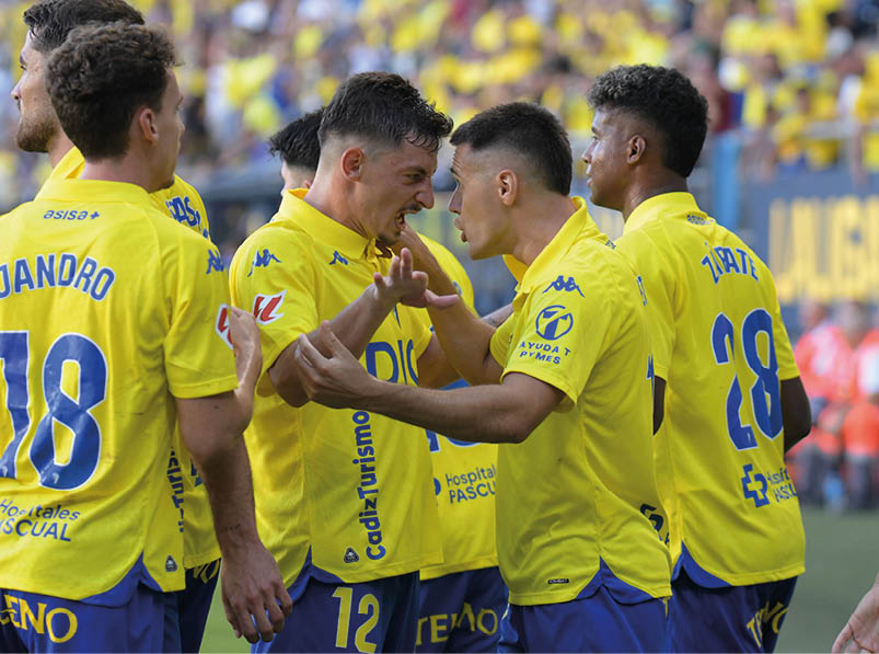 People wearing yellow sports jerseys are celebrating energetically on a soccer field with a crowd in the background. Descripci n generada con IA