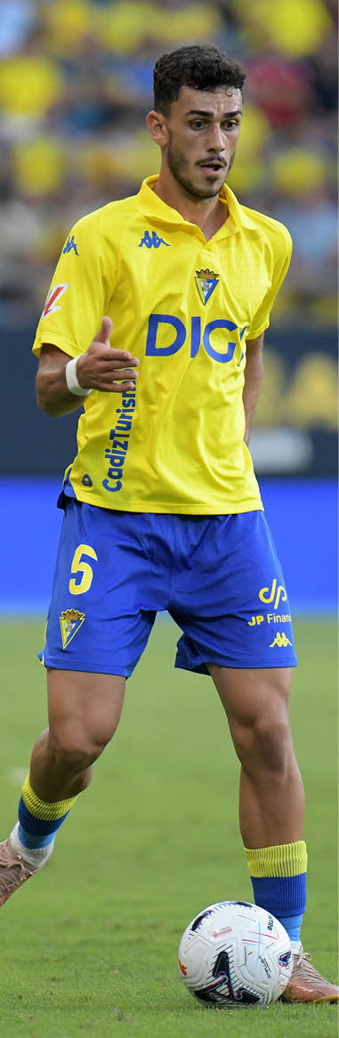 A person wearing a yellow and blue soccer uniform is standing on a grass field with one foot on a white soccer ball. Descripci n generada con IA