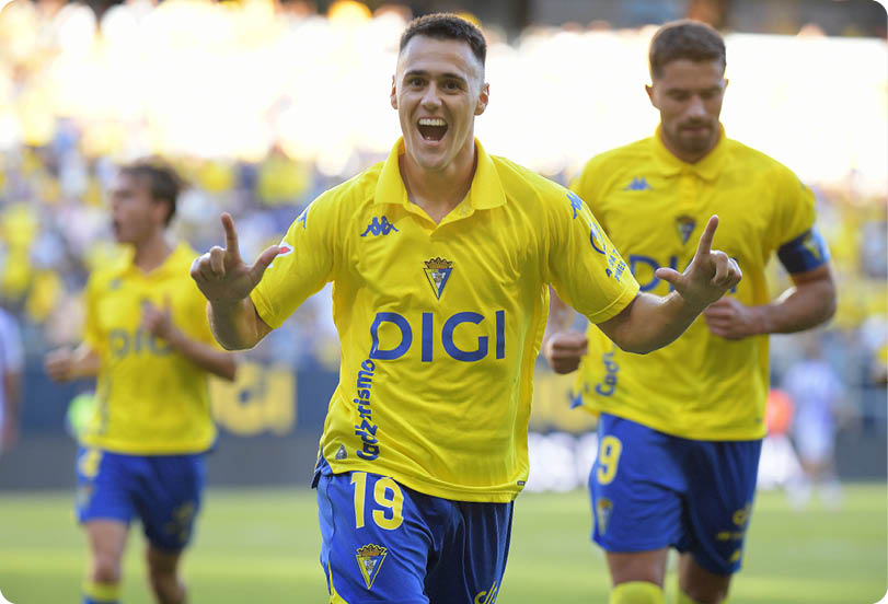 People wearing yellow and blue sports uniforms are celebrating on a soccer field, with one person in the foreground smiling and raising their hands. Descripci n generada con IA
