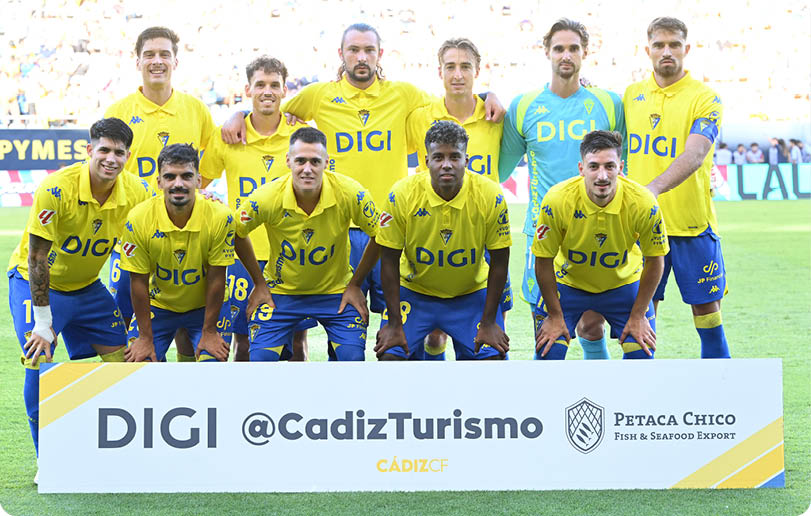 A group of people wearing yellow sports jerseys and blue shorts pose together on a soccer field behind a sign with the text DIGI @CadizTurismo and Petaca Chico Fish & Seafood Export. Descripci n generada con IA
