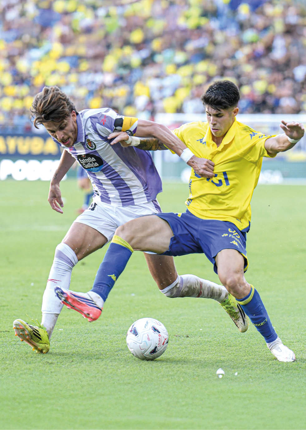 Two adults wearing sports uniforms are competing to kick a soccer ball on a grassy field with a blurred crowd in the background. Descripci n generada con IA