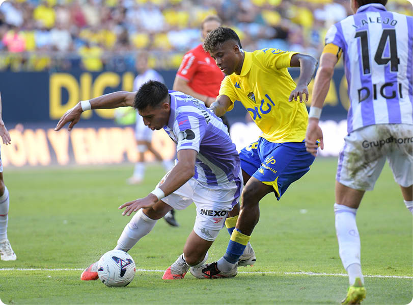 Two people in sports uniforms compete for a soccer ball on a grassy field during a match with a crowd in the background. Descripci n generada con IA