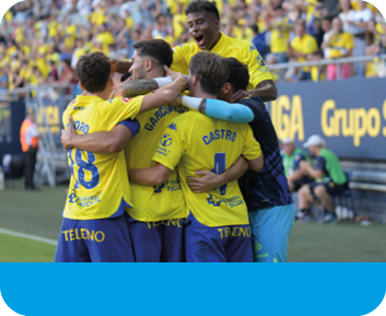 Un grupo de personas con uniformes deportivos amarillos y azules se abrazan celebrando en un campo de f tbol con p blico al fondo. Descripci n generada con IA