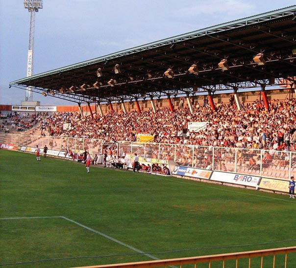 Multitud de personas reunidas en un estadio al aire libre con estructura met lica y techo cubierto. Descripci n generada con IA