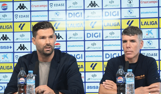 Dos personas sentadas frente a micr fonos en una conferencia de prensa con un fondo lleno de logotipos de patrocinadores deportivos. Descripci n generada con IA
