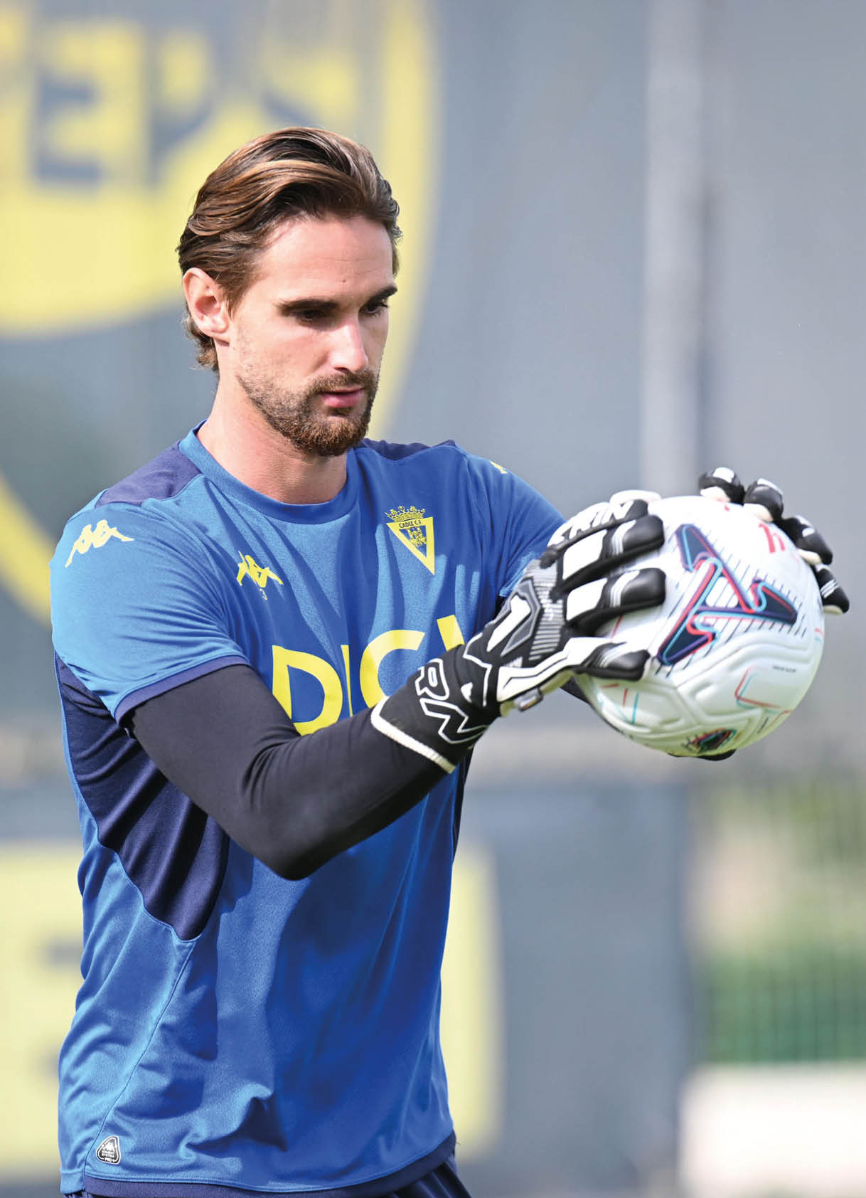 Una persona con camiseta azul y guantes sostiene un bal n de f tbol mientras parece concentrada en un entrenamiento al aire libre. Descripci n generada con IA