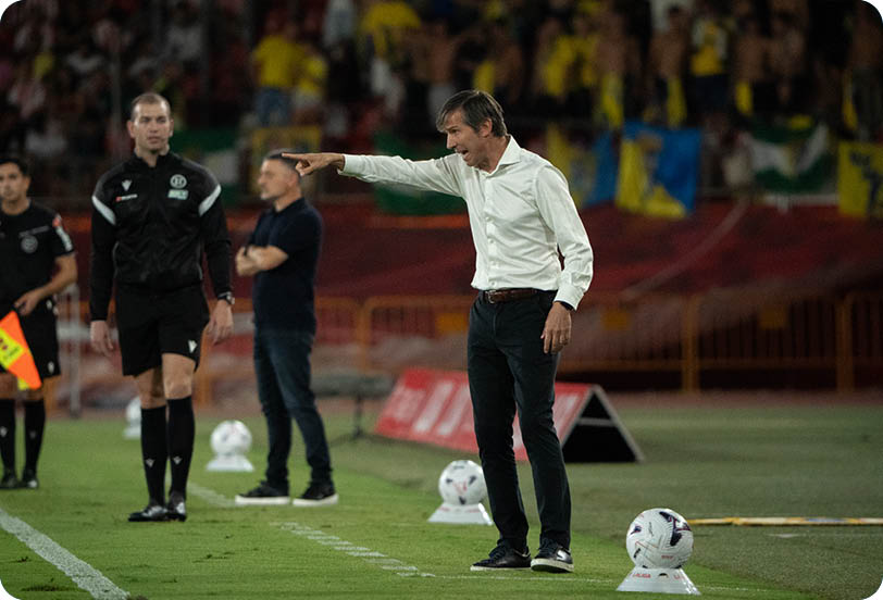 A person in a white shirt and dark pants is pointing while standing on the sidelines of a soccer field with two referees and another person in the background. Descripci n generada con IA