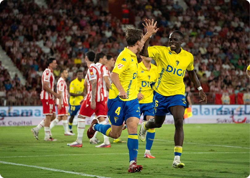 Two people in yellow and blue sports uniforms are celebrating with a high-five on a soccer field while other players in red and white uniforms walk in the background. Descripci n generada con IA