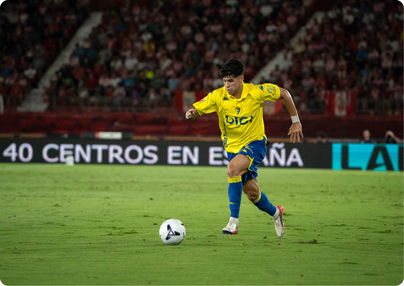 A person wearing a yellow and blue soccer uniform is running on a grass field while controlling a soccer ball during a game. Descripci n generada con IA