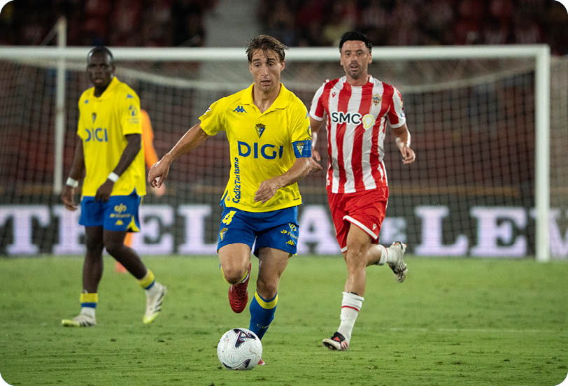 People in yellow and blue sports uniforms and a person in a red and white striped sports uniform are playing soccer on a grass field near a goal. Descripci n generada con IA