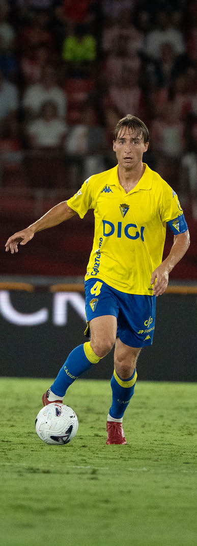 A person wearing a yellow and blue sports uniform is running on a grass field while controlling a soccer ball. Descripci n generada con IA