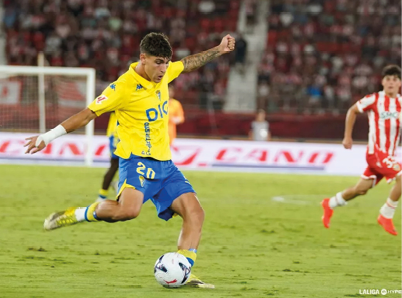 People wearing yellow sports jerseys are celebrating energetically on a soccer field with a crowd in the background. Descripci n generada con IA