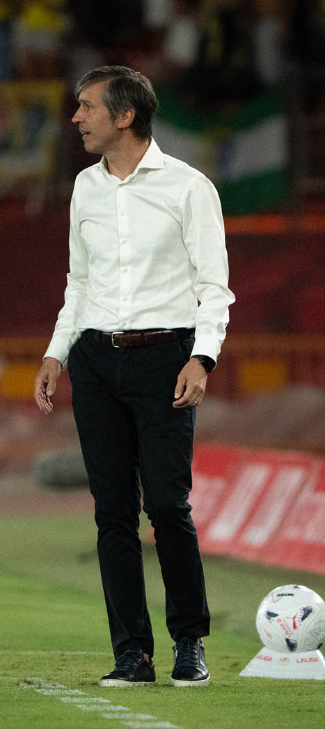 A person wearing a white button-up shirt and black pants stands on a grassy field near a soccer ball. Descripci n generada con IA