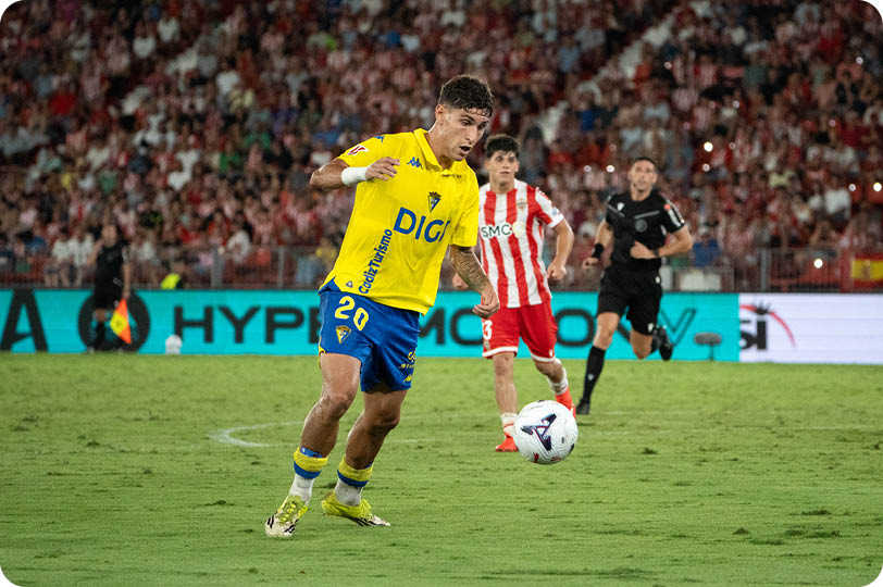People in yellow and blue sports uniforms and a person in a red and white striped sports uniform are playing soccer on a grass field near a goal. Descripci n generada con IA
