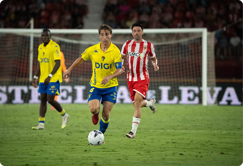Three people in sports uniforms are playing soccer on a grass field with one person in a yellow jersey running toward the ball while another in a red and white striped jersey follows closely behind. Descripci n generada con IA