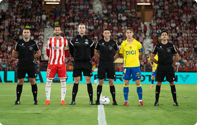 Five adults stand on a soccer field near the center line, with two wearing soccer uniforms and three wearing black referee outfits, and a crowd visible in the background. Descripci n generada con IA