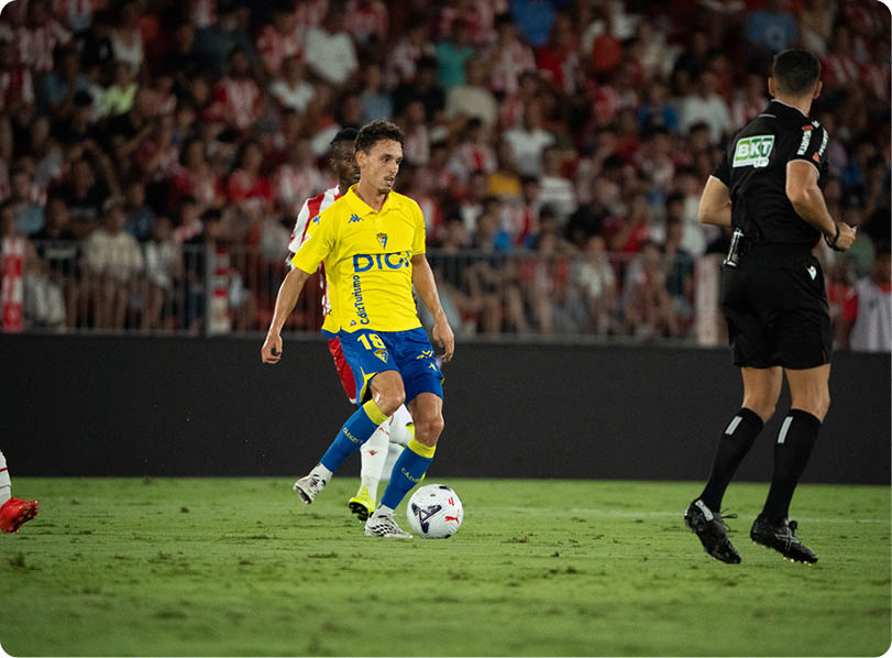 A person in a yellow and blue sports uniform is controlling a soccer ball on a grass field while another person in a black uniform and a person in a red and white uniform are nearby with a crowd in the background. Descripci n generada con IA