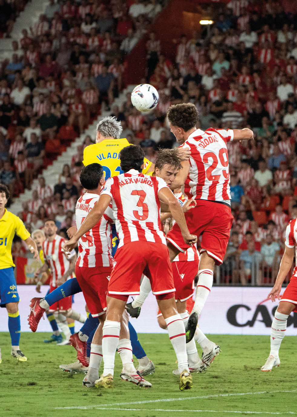 People in red and white striped sports uniforms and others in yellow and blue uniforms are actively competing for a soccer ball in a crowded stadium. Descripci n generada con IA