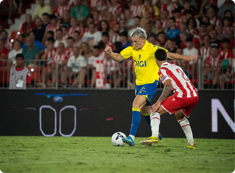Two adults in sports uniforms compete for a soccer ball on a grass field with a crowd watching in the background. Descripci n generada con IA