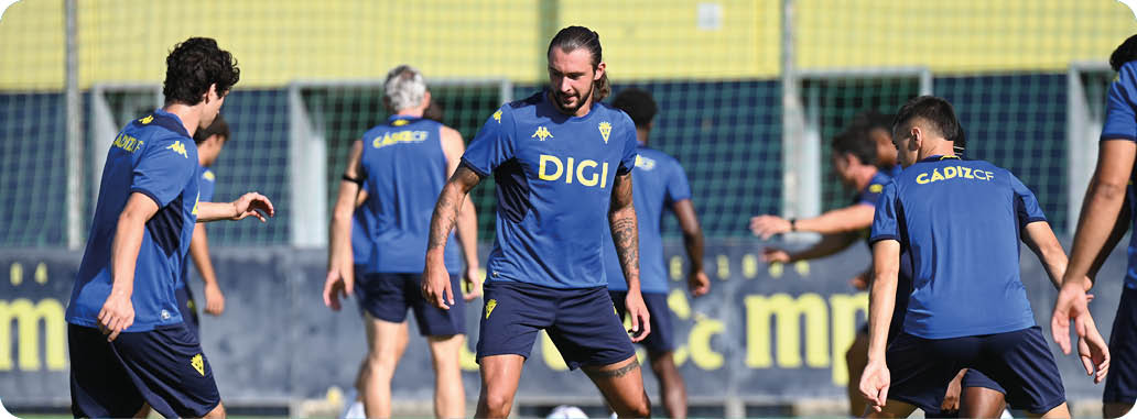 Un grupo de adultos con camisetas deportivas azules entrenan en un campo de f tbol con una porter a y una cerca al fondo. Descripci n generada con IA