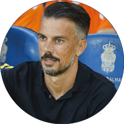 Persona con cabello corto y barba canosa vistiendo una camisa negra sentada en un estadio deportivo con personas vestidas de naranja al fondo. Descripci n generada con IA