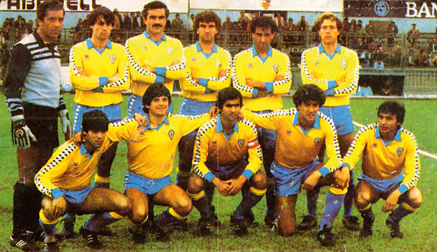 Un grupo de personas con uniformes deportivos amarillos y azules posan en un campo de f tbol, algunas de pie con los brazos cruzados y otras agachadas. Descripci n generada con IA