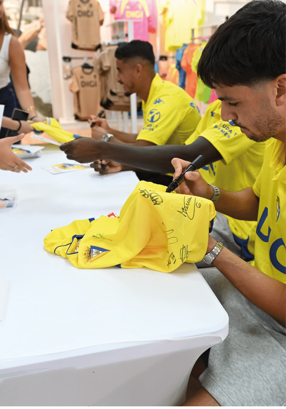 Personas con camisetas amarillas est n firmando camisetas del mismo color en una mesa blanca dentro de una tienda o evento. Descripci n generada con IA