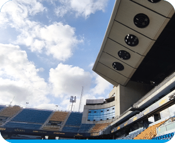 Estadio vac o con gradas de colores azul y amarillo bajo un cielo parcialmente nublado y una estructura met lica con luces en el techo. Descripci n generada con IA