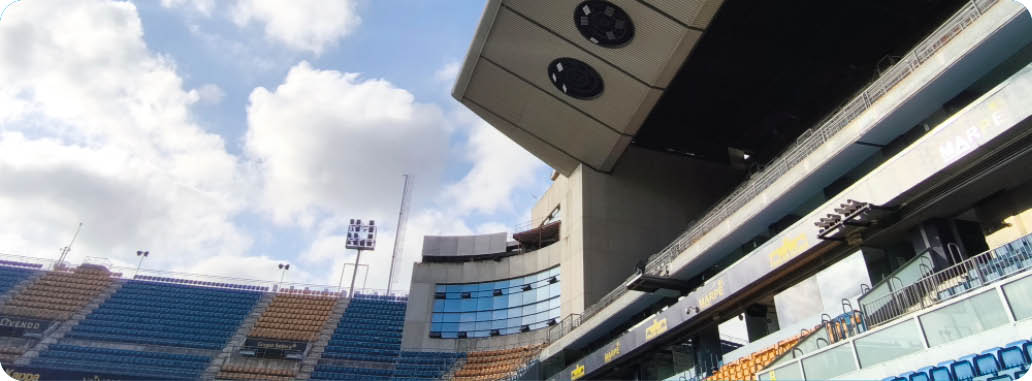 Estadio vac o con gradas de colores y una estructura grande con luces bajo un cielo parcialmente nublado. Descripci n generada con IA