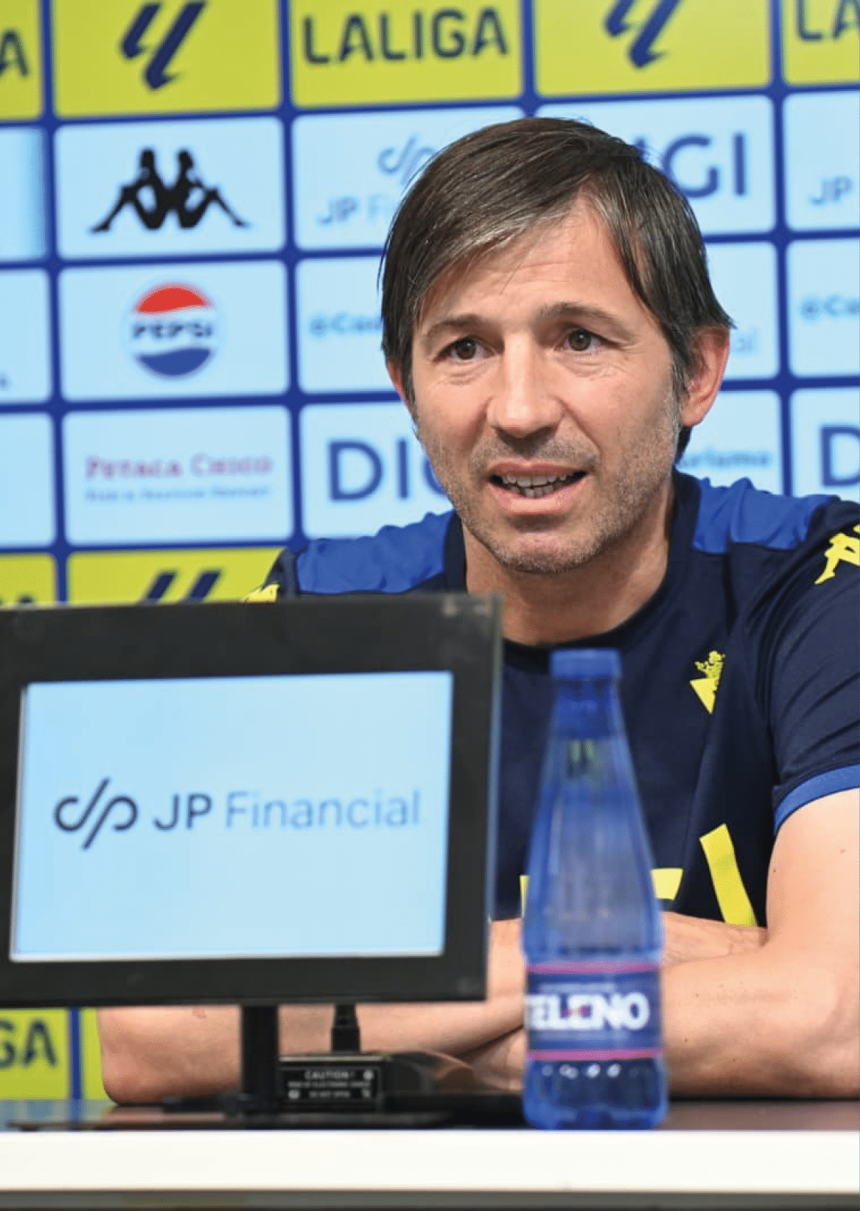Una persona con camiseta deportiva azul habla en una conferencia de prensa con un fondo lleno de logotipos y una botella de agua sobre la mesa. Descripci n generada con IA