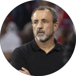 Una persona adulta con barba y cabello corto canoso viste una camiseta negra de manga larga y parece estar observando algo con expresi n seria en un entorno posiblemente deportivo. Descripci n generada con IA