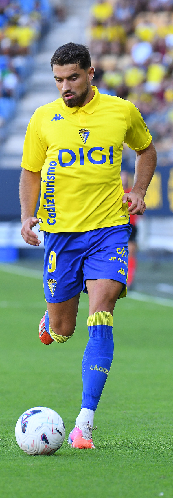 Persona con uniforme deportivo amarillo y azul controlando un bal n de f tbol en un campo. Descripci n generada con IA