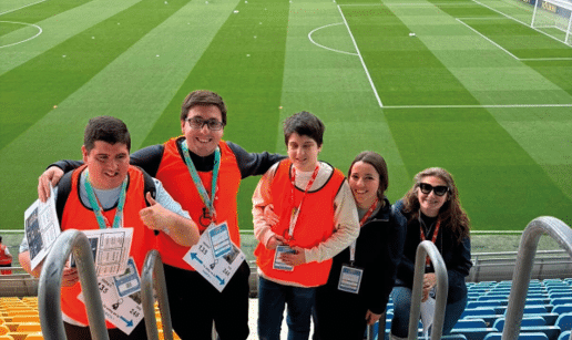 Cinco personas con chalecos naranjas y gafetes sonr en y posan juntas en las gradas de un estadio de f tbol con c sped verde. Descripci n generada con IA
