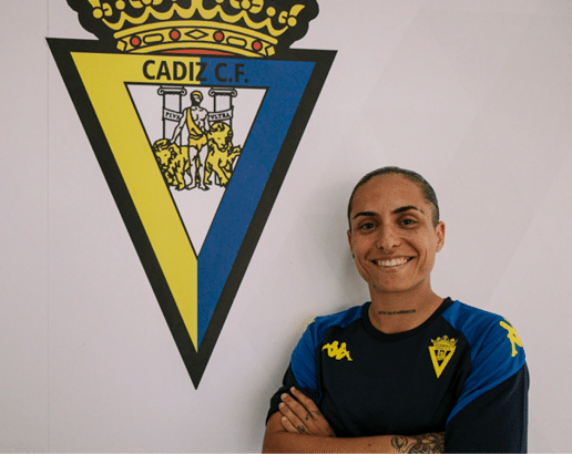 Persona sonriente con camiseta deportiva azul y negra con el escudo del C diz C.F. posando junto a un gran escudo del equipo en la pared. Descripci n generada con IA