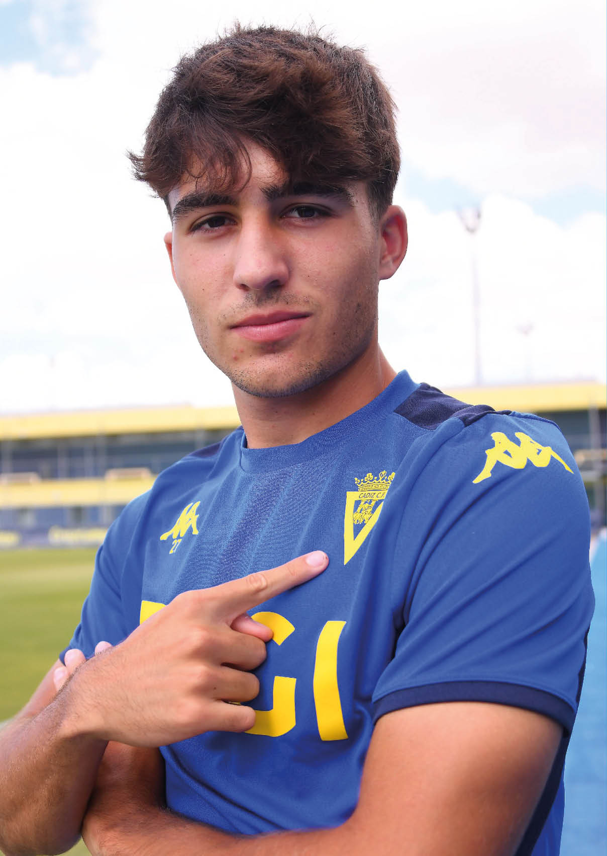 Persona joven con cabello corto y rizado lleva puesta una camiseta deportiva azul con detalles amarillos y se ala con el dedo ndice el escudo en el pecho mientras est  al aire libre en un campo deportivo.  Descripci n generada con IA