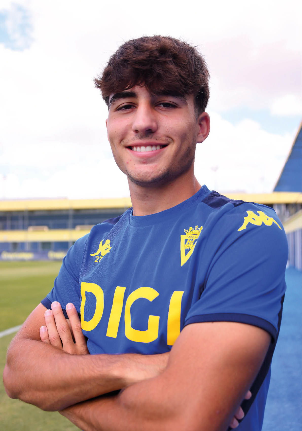 Persona joven con cabello corto y rizado sonriente con los brazos cruzados vistiendo una camiseta deportiva azul con detalles amarillos en un campo de f tbol. Descripci n generada con IA