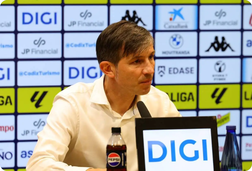 A person with short hair wearing a dark polo shirt sits at a table with a bottle of Pepsi and a water bottle in front of a backdrop covered with various sponsor logos. Descripci n generada con IA