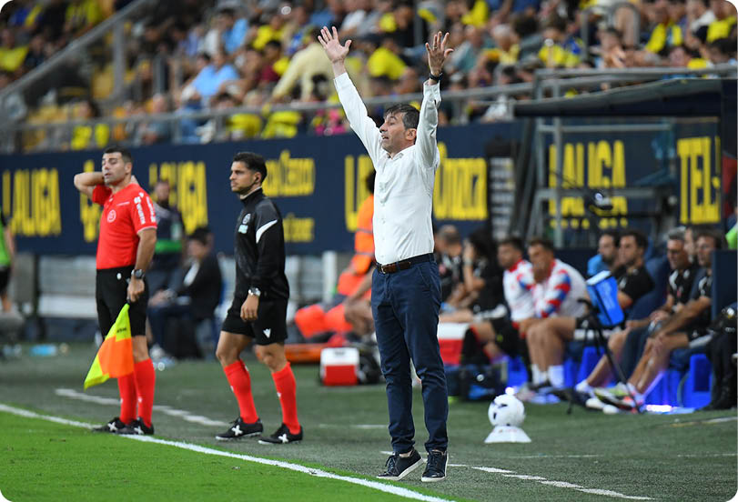 A person in a white shirt and dark pants is standing on the sidelines of a soccer field with arms raised while two referees in black and red uniforms stand nearby and a group of people sit on a bench in the background. Descripci n generada con IA