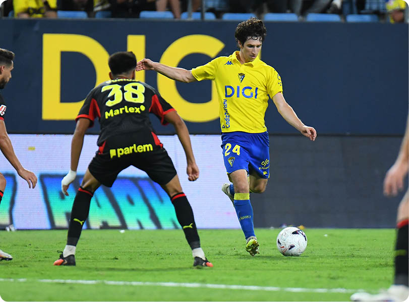 A person in a yellow and blue soccer uniform is about to kick a soccer ball while another person in a black and red uniform stands in a defensive position on a soccer field. Descripci n generada con IA