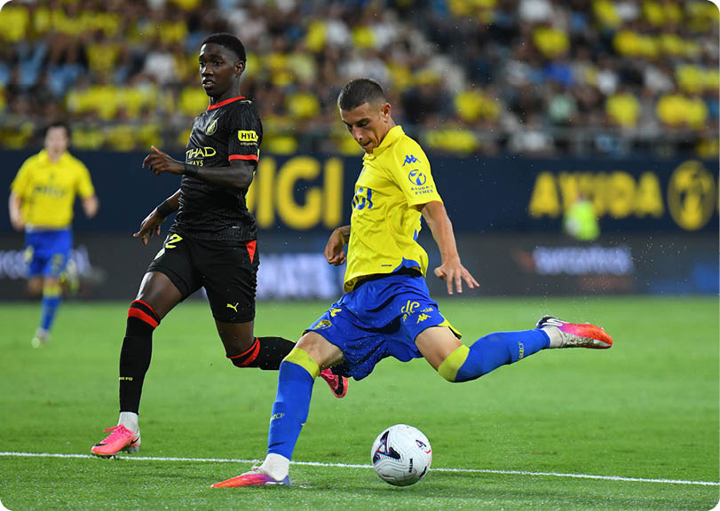 Two people in sports uniforms are actively playing soccer on a field with one person kicking the ball while the other is running nearby. Descripci n generada con IA